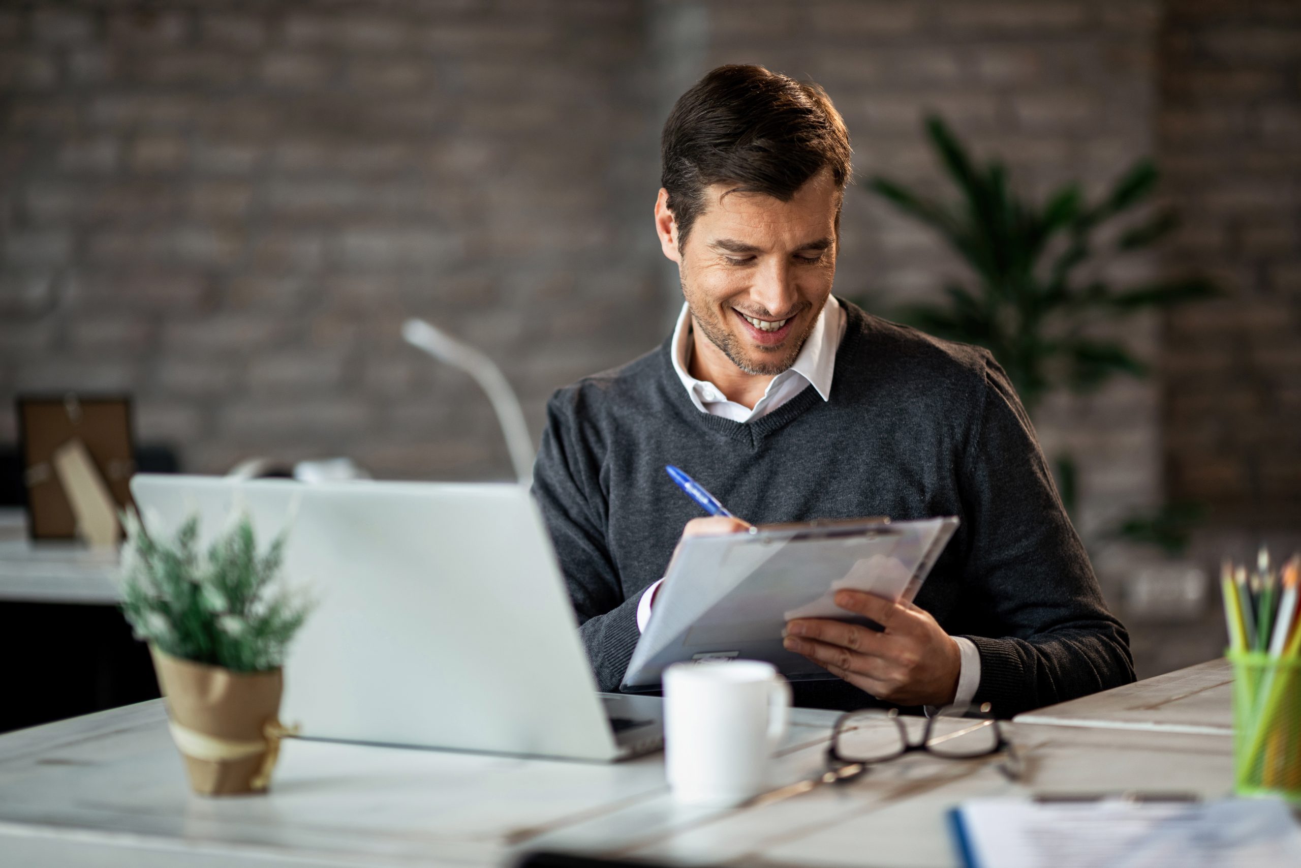 Smiling entrepreneur writing business report while working on laptop in the office.